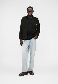 Jachetă neagră din catifea cu nasturi, asortată cu un tricou negru și blugi deschiși la culoare. Pantofi maro cu un finisaj neted completează ținuta.