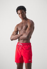 Pantaloni da bagno rossi realizzati in tessuto leggero, con vita elastica e cordoncino bianco, e un logo sulla parte inferiore della gamba sinistra.