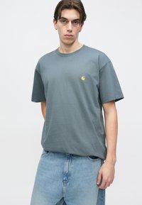 Νέος άνδρας με καστανά μαλλιά που φορά γκρι μπλουζάκι Carhartt και ανοιχτόχρωμο τζιν μπλε μπροστά σε απλό λευκό φόντο.