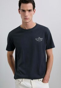 Camiseta de algodón gris oscuro con mangas cortas, cuello redondo y un pequeño gráfico blanco en el lado izquierdo del pecho. Combinada con pantalones de color claro.