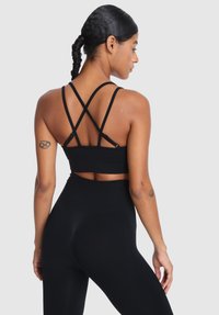 Žena s opletenými vlasmi, oblečená v čiernej športovej podprsenke s prekrývajúcimi sa pásikmi a v legínach s vysokým pásom, ukazujúc tetovania na horných ramenách, stojí chrbtom.