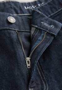 Donkerblauwe baggy jeans met een metalen rits, zichtbare stiksels en een gemerkte knoop in de tailleband. De textuur lijkt op denim.