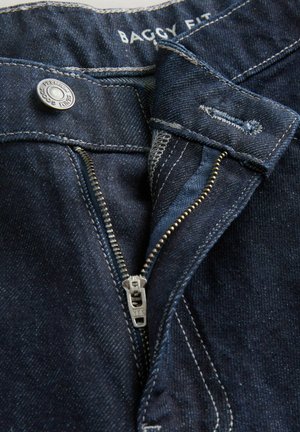 Dark blue baggy fit jeans with a metal zipper, visible stitching, and a branded button at the waistband.  Texture appears denim.