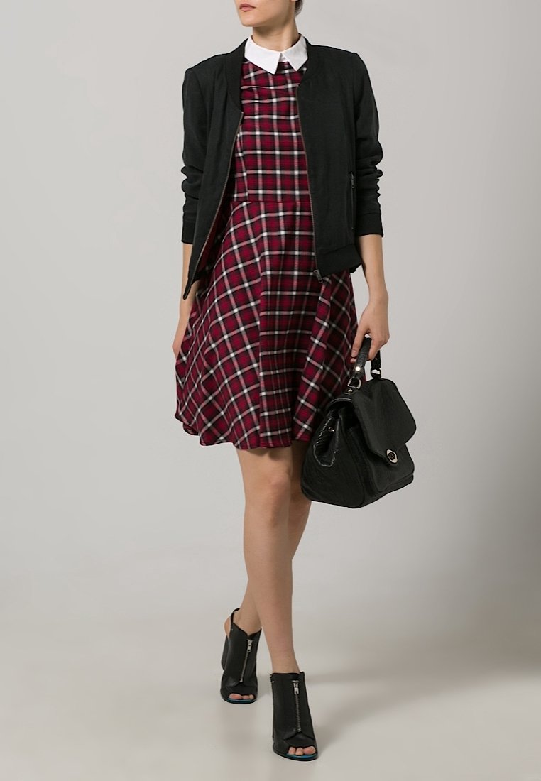 Černá bomber bunda přes červenou kostkovanou šaty s bílým límcem, doplněná černými otevřenými kotníkovými botami a černou kabelkou.