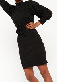 Musta mekko korkealla pääntiellä, puffitakeilla ja vyötärölle sidotulla vyöllä. Valmistettu tekstuurista kankaasta, jossa on hienovarainen kiilto. Keskimmäiseen reiteen ulottuva pituus.