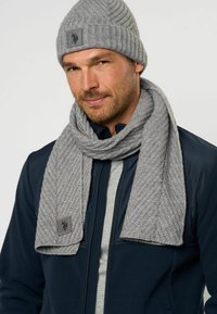 Grå ribbet strikket hue og matchende tørklæde, tekstureret med et houndstooth-mønster. Tørklædet har et lille logo-lander. Båret over en marineblå jakke.