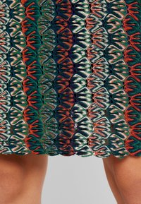 Szydełkowa spódnica z falbanistym wzorem. Oferuje zielone, pomarańczowe i kremowe wzory w interlokach, tworząc teksturowany efekt warstwowy.