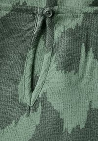 Vihreä kuvioitu kangas, jossa on tekstuuri; siinä on nappikiinnitys ja pieni aukko. Suunnittelussa on abstrakteja muotoja eri sävyissä.