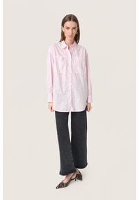 Camicia button-up rosa chiaro con maniche lunghe e due taschini sul petto, abbinata a jeans a gamba larga grigio scuro e scarpe a strisce bianche e nere.