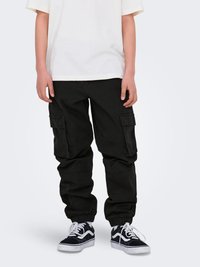 Črne cargo hlače z elastičnim pasom in manšetami, ki vsebujejo več stranskih žepov. Nosi se jih z belo majico in črnimi supergami z belimi podrobnostmi.