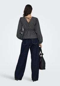 Femme vue de dos portant un chemisier à carreaux sombre avec un détail de nœud dans le dos et un jean large sombre, tenant un sac à main noir dans sa main droite.