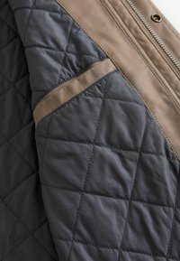 Chaqueta acolchada con un cuerpo en gris oscuro, acentos de cuero color beige y una franja diagonal beige a lo largo del costado. Materiales suaves y texturizados.