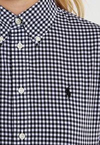 Black and white gingham check button-up shirt, featuring a pointed collar, pearl buttons, and a black embroidered logo on the left chest.