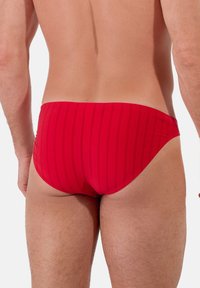 Czerwone kąpielówki z pionowymi prążkami, minimalna osłona, gładka tekstura i jednolita design pasa. Widok z tyłu ukazuje dopasowanie i styl.