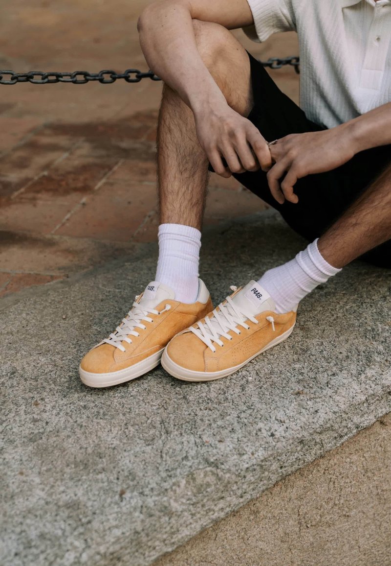 Personne assise sur un banc en pierre portant des chaussettes blanches, des baskets jaunes, un short noir et une chemise blanche à manches courtes, les mains jointes près des genoux.
