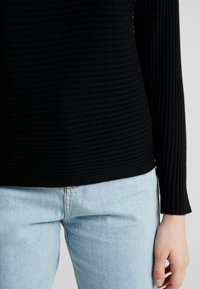 Černý žebrovaný top s dlouhým rukávem z texturované látky, kombinovaný se světle modrými džínami. Top má přiléhavý střih a kulatý výstřih.