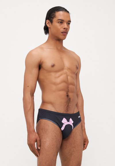 Shorts de bain noirs avec un détail en forme de nœud rose à l'avant, en matériau lisse, avec une taille élastique et une coupe ajustée.
