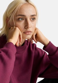 Sudadera burdeos con cuello alto y puños acanalados; el modelo tiene el cabello corto y rubio y descansa las manos sobre las mejillas. Fondo neutral.