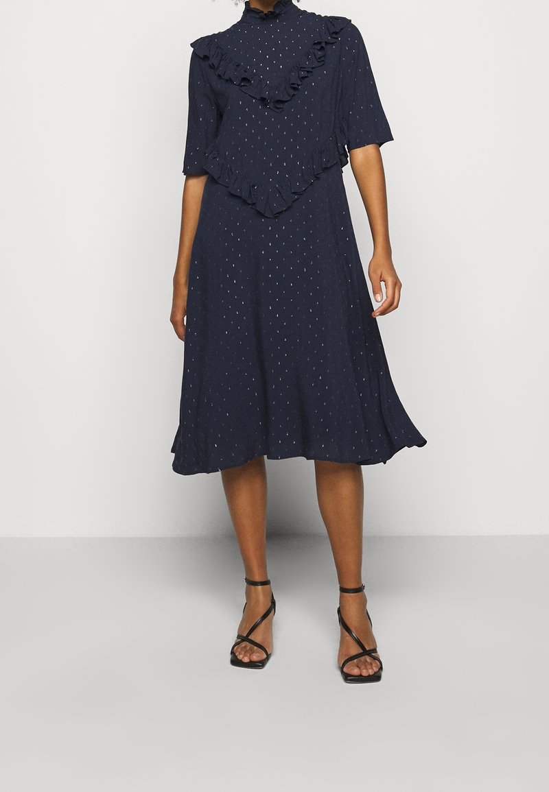 Femme portant une robe midi bleu marine à pois, avec des détails volantés sur la poitrine et les manches, et des sandales noires à talons hauts à brides.