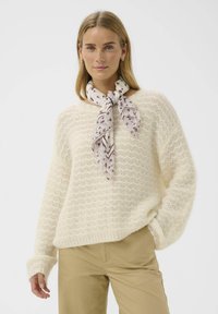 Pull en tricot couleur crème avec un motif ondulé, associé à une écharpe légère à motifs nouée au cou. Texture douce et coupe décontractée.