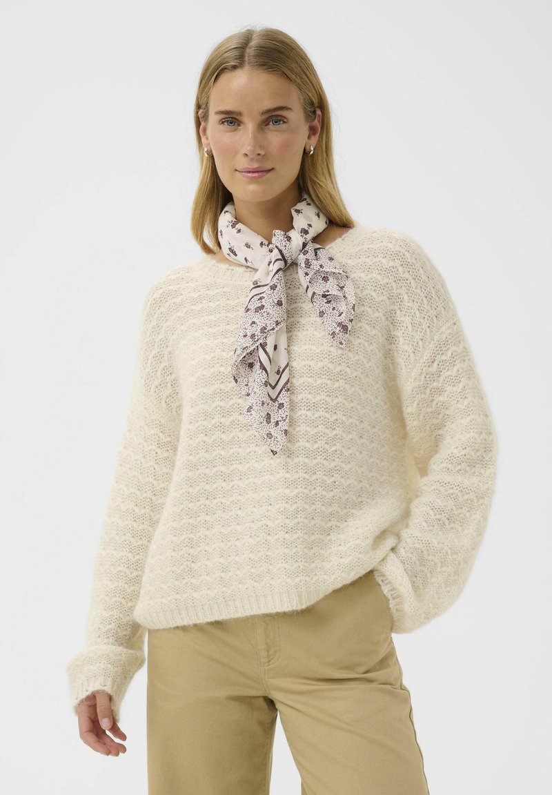 Pull en tricot couleur crème avec un motif ondulé, associé à une écharpe légère à motifs nouée au cou. Texture douce et coupe décontractée.