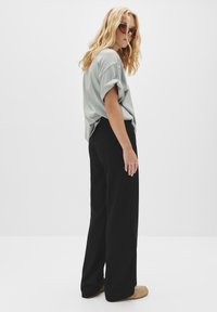 Blonde vrouw in opgerolde lichtgrijze T-shirt, zwarte wijde broek, beige schoenen en donkere zonnebril, staand tegen een effen witte achtergrond.