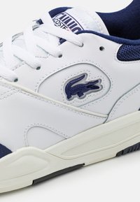 Fehér bőr sportcipő tengerészkék kiegészítőkkel, textúrázott fehér talppal, fűzőzárral és tengerészkék Lacoste logóval a nyomott részén.