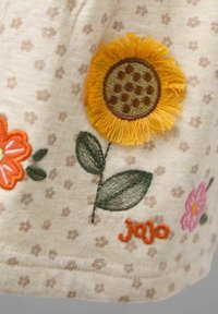 Šviesiai smėlinė medžiaga su rožinėmis gėlių raštais ir spalvingais siuvinėjimais, įskaitant saulėgrąžą. "Jojo" yra siuvinėtas oranžine spalva.