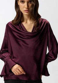 Burgundijska satenska bluzica s okruglim izrezom, dugim rukavima i rukavima u obliku zvona. Glatka tekstura i elegantan pad.
