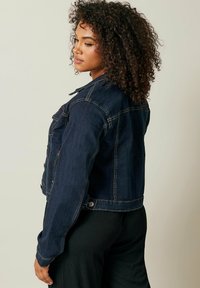 Tumma sininen farkkutakki, jossa on lyhennetty malli, napitukset ja klassinen kaulus. Siinä on näkyvät ompeleet ja pitkät hihat.