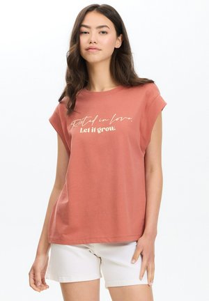 Joven mujer con cabello largo y oscuro, que lleva una camiseta sin mangas de color óxido con texto y pantalones cortos blancos sobre un fondo blanco.
