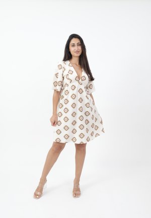 Weißes Kleid mit V-Ausschnitt und Puffärmeln, ausgestattet mit einem braunen geometrischen Muster. Kombiniert mit klaren Absatzsandalen.