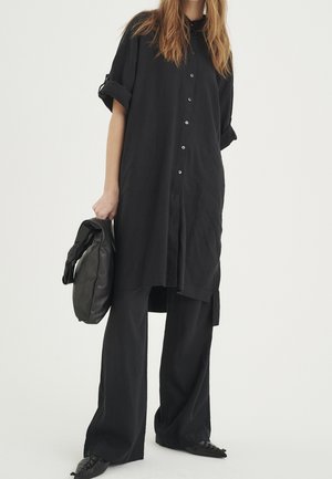 Chemise noire surdimensionnée à boutons et manches retroussées, associée à un pantalon noir à jambes larges et un sac noir à texture douce. Les chaussures présentent un bout pointu.