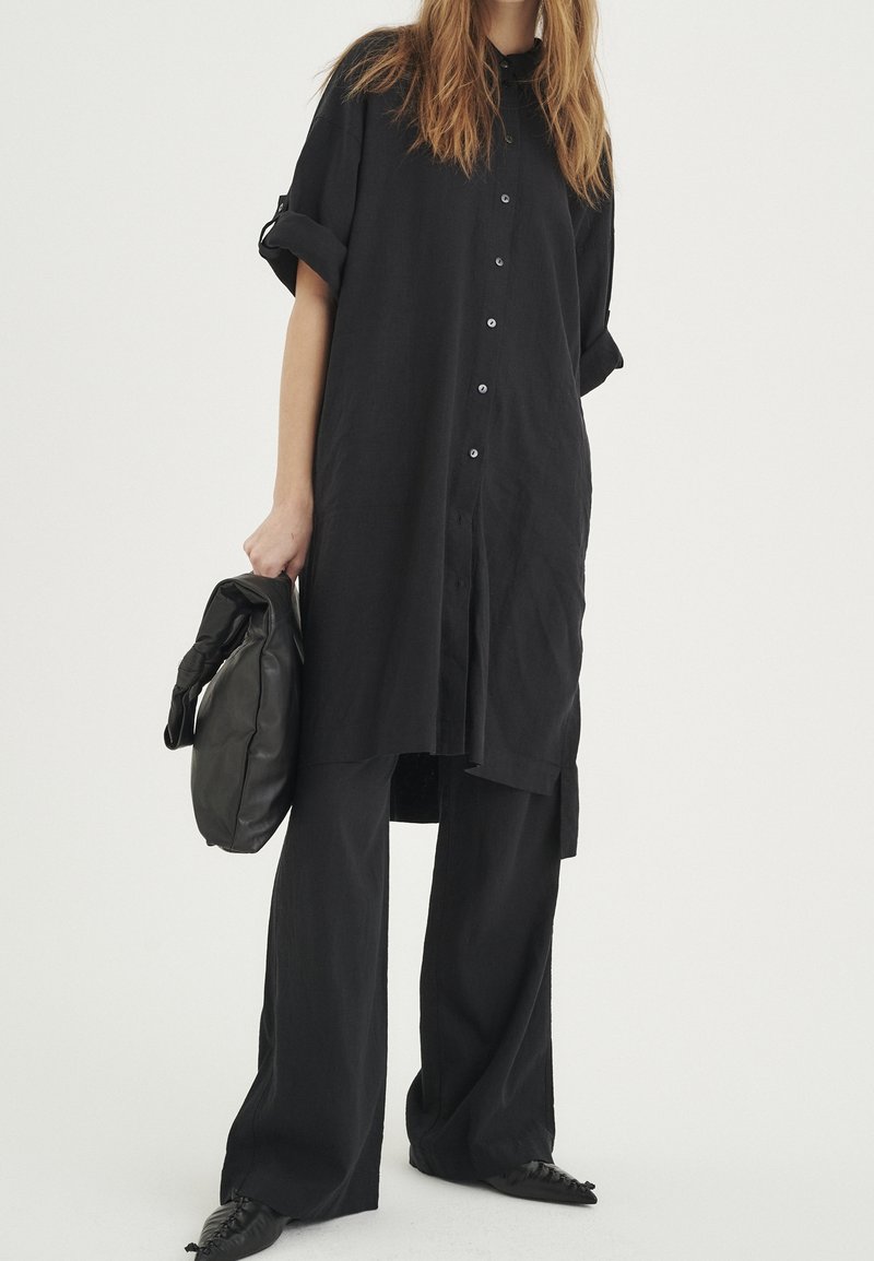Chemise noire surdimensionnée à boutons et manches retroussées, associée à un pantalon noir à jambes larges et un sac noir à texture douce. Les chaussures présentent un bout pointu.