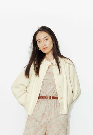 Jeune femme aux cheveux longs et foncés portant une veste crème sur une robe à fleurs cintrée avec une ceinture marron, debout devant un fond blanc.