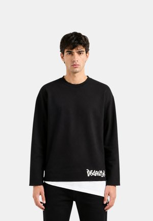 Giovane uomo con capelli scuri che indossa una maglia nera a maniche lunghe con la scritta bianca “DISCLAIMER” vicino all'orlo e una camicia bianca sotto.
