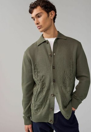 Olive green cardigan with textured floral embroidery on the front, buttons, and ribbed cuffs. White t-shirt underneath, casual fit.