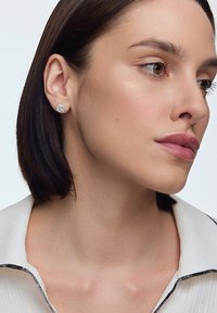 Srebrne kolczyki w kształcie studs z kwadratową, teksturowaną powierzchnią i małymi przezroczystymi kamieniami, noszone przez modelkę z krótkimi włosami.