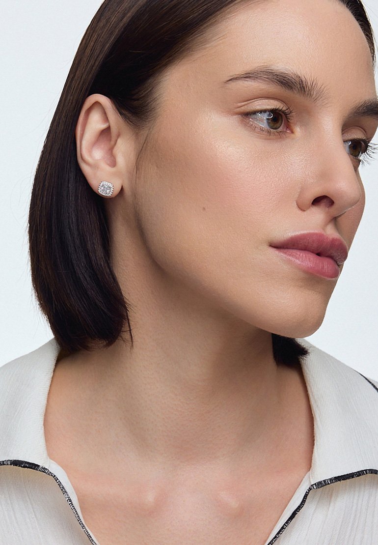 Srebrne kolczyki w kształcie studs z kwadratową, teksturowaną powierzchnią i małymi przezroczystymi kamieniami, noszone przez modelkę z krótkimi włosami.