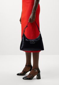 Sac en cuir noir à bandoulière avec une forme courbée, présentant une finition texturée et un accent de marque. Porté avec une robe rouge ajustée et des talons noirs.