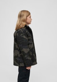 Tarnjacke mit dunklen grünen und schwarzen Mustern, knopfverschlossen vorne und seitlichen Taschen, getragen von einer Person mit hellbraunen Haaren.