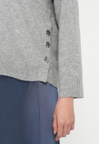 Pulover tricotat de culoare gri deschis, cu manșete și tiv rib înfășurat, având trei nasturi decorativi pe lateral, asortat cu o țesătură de culoare bleumarin.