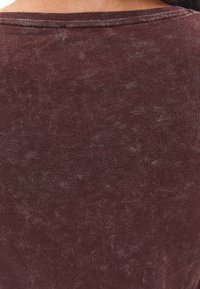 Braunes, strukturiertes Baumwolloberteil mit Rundhalskragen, das eine leicht melierte Optik und eine taillierte Passform aufweist. Schlichte und glatte Oberfläche.