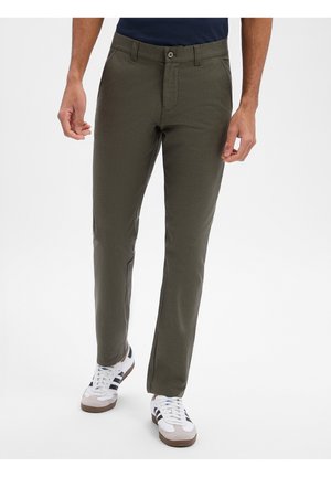Man draagt olijfgroene slim-fit broek en witte sneakers met zwarte strepen, lopend tegen een effen witte achtergrond.