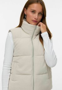 Beige gewatteerde vest met hoge kraag, ritssluiting en zijzakken. Gemaakt van ribbelsweefsel, met subtiele horizontale stikdetails.