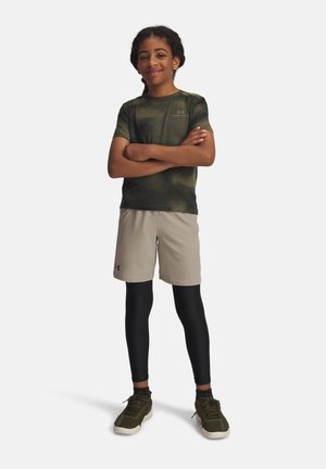 Garçon debout, bras croisés, portant un t-shirt camouflage vert, un short beige sur des leggings noirs et des baskets vert olive sur fond blanc.