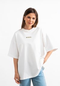 Witte oversized t-shirt met "MIRONS" geborduurd in goud, ronde hals, korte mouwen en een relaxte pasvorm, gecombineerd met blauwe denim jeans.
