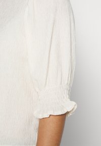 Manica di una blusa bianca texturizzata con polsino elastico e bordo arricciato sul braccio di una persona, su uno sfondo neutro.