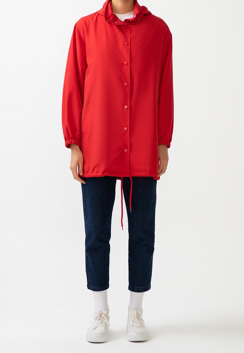 Rød vandtæt jakke med hætte, trykknapper og elastiske manchetter. Bæret over mørke jeans og hvide sneakers. Enkel, funktionel design.