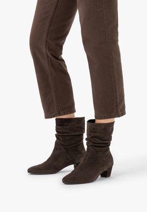 Bottes molles en daim brun foncé montant jusqu'au milieu du mollet, portées avec un pantalon marron assorti coupé au-dessus de la cheville sur un fond blanc.